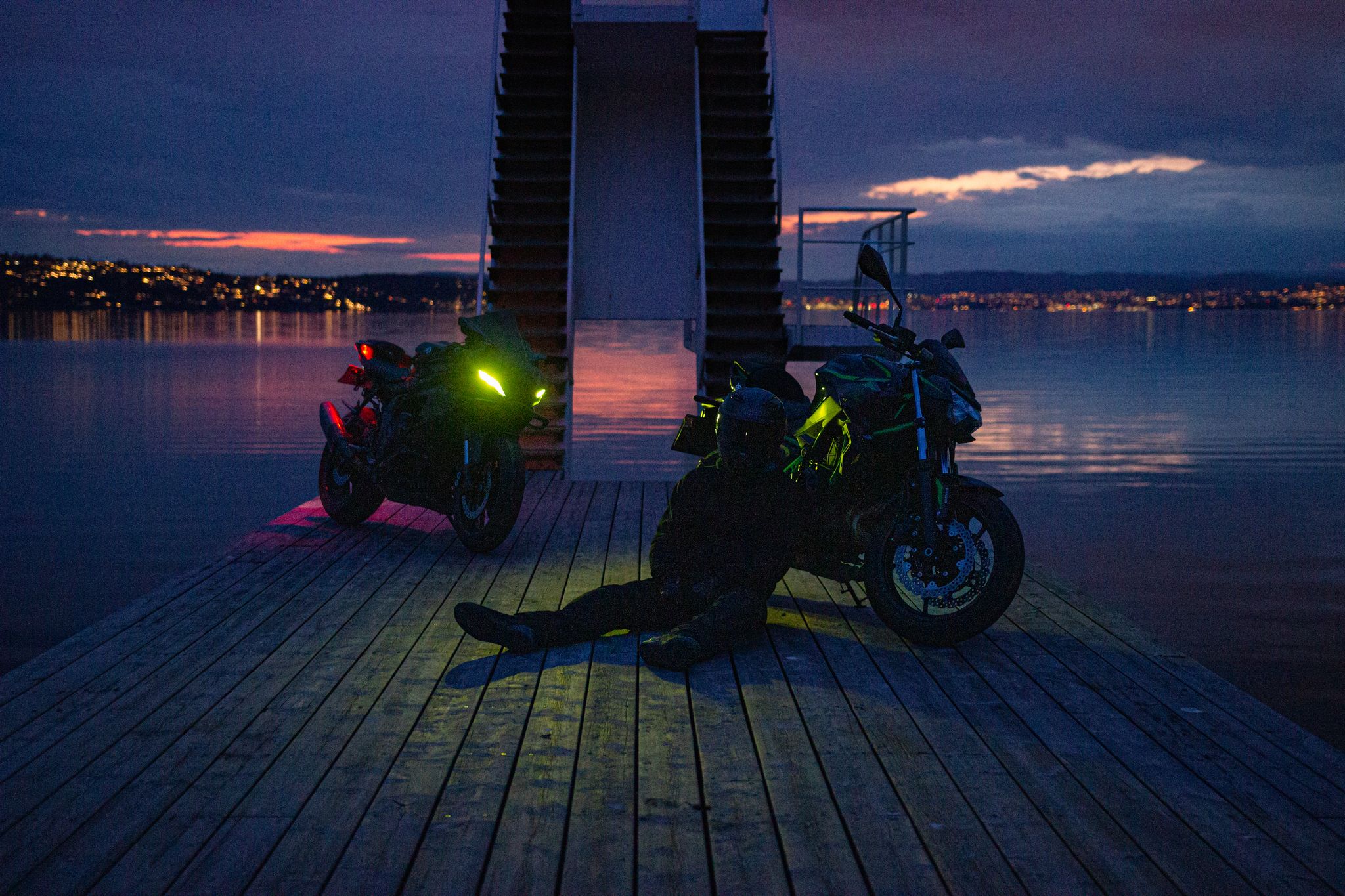 To motorsykler på brygga i blåtimen, lysene på, speilinger i vannet